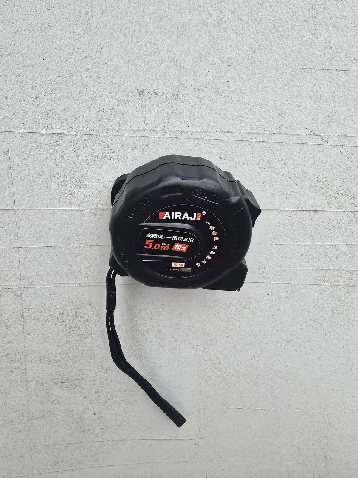 A 5m tape measure hanging on the wall-mounted holder, wrist strap loose below.