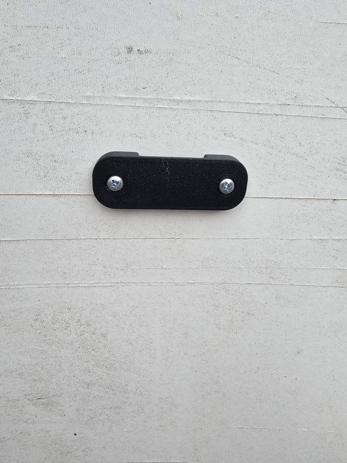 The black tape measure holder screwed to a painted timber wall with two screws.